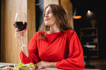 happy young woman holding glass with red wine and smiling near plate with salad in restaurant