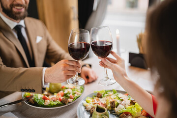 cropped view of couple clinking glasses with red wine near meal during celebration on valentines day