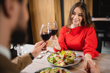 cheerful woman holding hands with boyfriend while clinking glasses of red wine