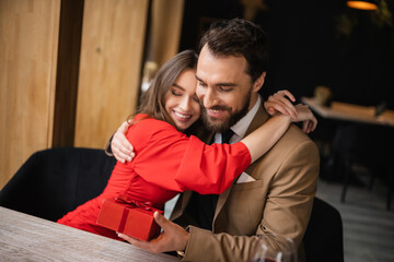 cheerful woman in red dress hugging bearded man in formal wear holding present on valentines day