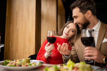 young woman and cheerful man holding glasses with red wine during celebration on valentines day