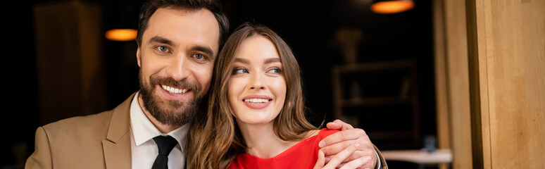 portrait of cheerful and bearded man looking at camera while hugging girlfriend, banner