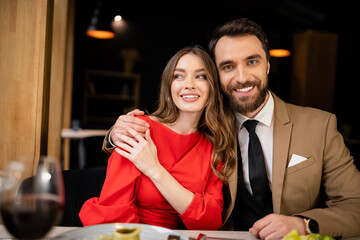 bearded man hugging cheerful young woman in red dress and looking at camera on valentines day