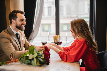 happy woman in red dress holding hands with man near bouquet of roses on valentines day