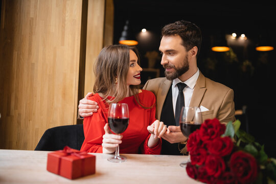 bearded man holding hand of cheerful woman with engagement ring on finger on valentines day