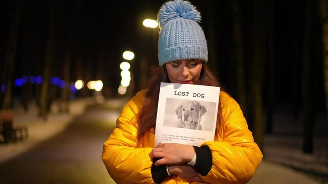 Frustrated young woman with lost dog announcement standing on winter night outdoors looking around. Portrait of sad depressed Caucasian lady searching pet on city street