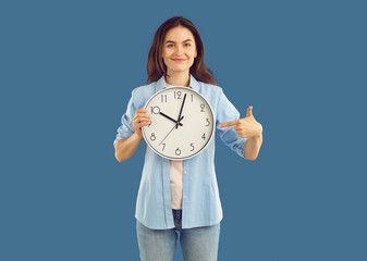 Portrait of beautiful happy young brunette holding round wall clock in one hand and pointing her...