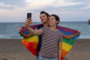 Beautiful gay young couple embraces and holds a rainbow flag. Happy couple taking selfie photo at the beach..