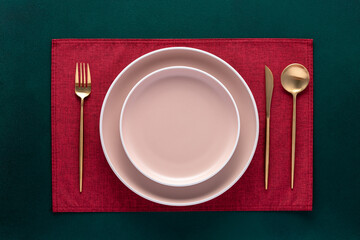 Empty plate with fork and knife on dark green table. Festive place setting with red napkin and gold...