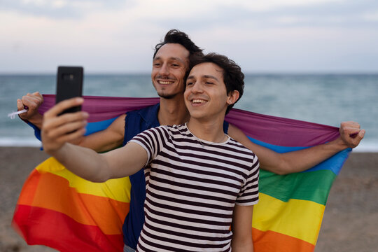 Beautiful gay young couple embraces and holds a rainbow flag. Happy couple taking selfie photo at the beach..
