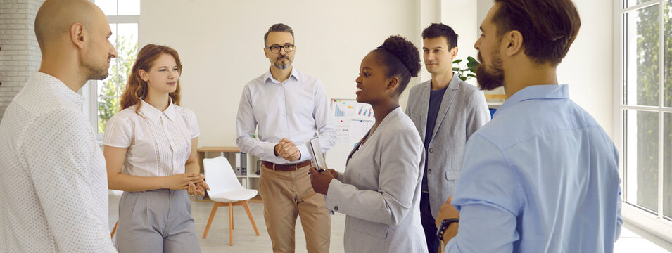 Woman manager gives assignments to her employees during meeting before start of working day. Men and women stand in circle and listen to their African American female leader. Panoramic view. Banner.