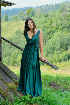 Pretty brunette girl standing at old house in the carpathians. Young romantic woman in long, green, stin dress on the mountainside.