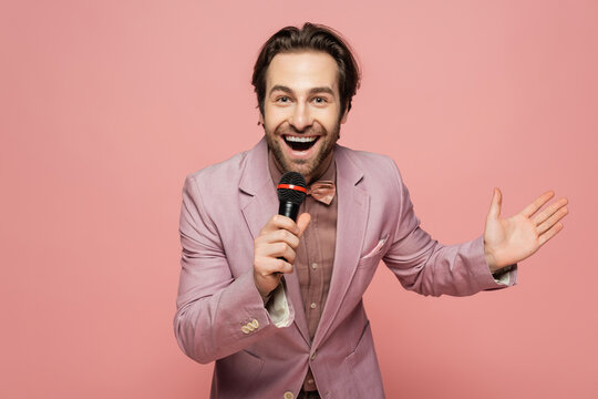 Cheerful Host Of Event Talking At Microphone And Waving Hand Isolated On Pink