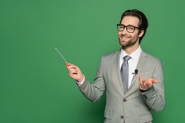 cheerful news anchor in suit and eyeglasses smiling while pointing with pencil on green background