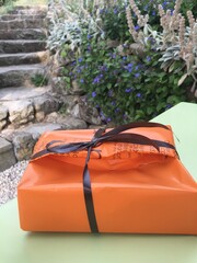 Ein Geschenk, dass in orangene Folie verpackt wurde liegt auf einem Tisch. Im Hintergrund sind...