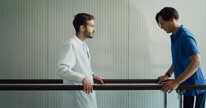 Young Male Doctor is Working With a Patient After Serious Injury. Professional Physician Helping Man to Recover After Unfortunate Accident. Walking Using Rehabilitation Parallel Bars As Support.