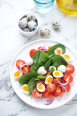 Salad with fresh spinach, red cherry tomatoes and boiled quail eggs, vertical shot on a white marble background, elevated view