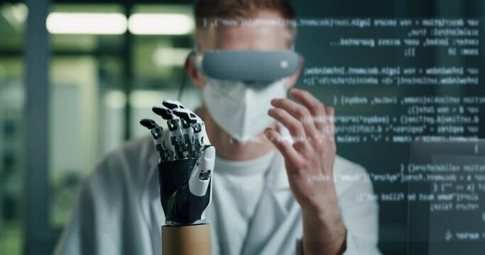 Young Biomedical Engineer is Wearing Virtual Reality Goggles and Controlling Remotely Modern Bionic Limb Prosthesis. Skilled Professional Programming Artificial Palm. Lines of Code Running on Screen.