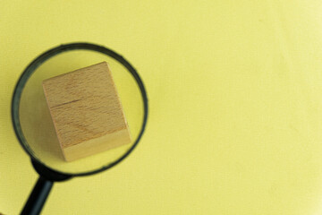 Photo of magnifying glass and focus on blank wooden cube on left side over yellow background with copyspace for put your text or logo.,Flat lay top view mock-up item concept.