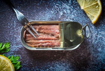 Canned anchovies open on a dark blue table with a fork, lemon and parsley. Ready for eat. 