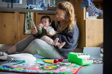 Mother and baby playing with their home clothes in the kitchen of the messy house in the morning, looking at the notifications on the phone.Daily lifestyle, mom life concept. Real home environment.