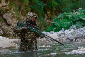 A military man or airsoft player in a camouflage suit sneaking the river and aims from a sniper rifle to the side or to target.