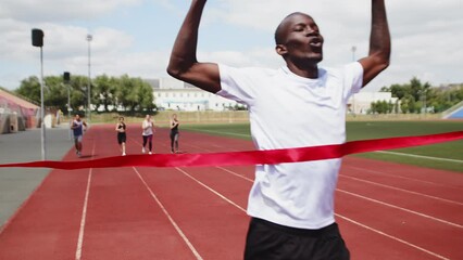 Young fit black man crossing finish line and tearing red ribbon finishing faster than rivals in running competition at the outdoor stadium in the summer