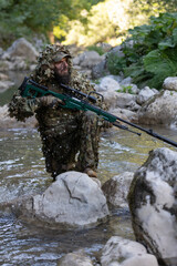 A military man or airsoft player in a camouflage suit sneaking the river and aims from a sniper rifle to the side or to target.