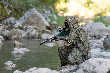 A military man or airsoft player in a camouflage suit sneaking the river and aims from a sniper...