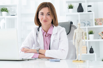 Portrait photo of young and beautiful smiling female doctor in white medical gown sitting and working on laptop in the office of the modern clinic. Online consultation and distant cure concept