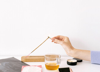 woman hand lighting an incense stick on the table preparing for practice with cup of tea and candle light, mental relaxation cover, copy space