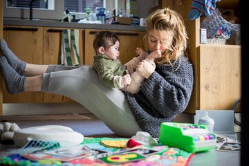 The young mother, kissing the feet of her baby on her knees, spends time playing with her baby in the messy kitchen environment.
Daily lifestyle. Intimate real authentic moment. - obrazy, fototapety, plakaty