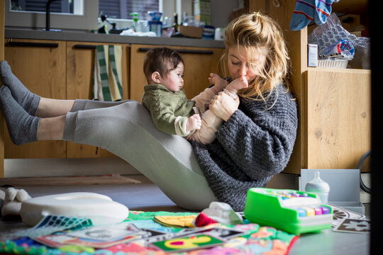 The young mother, kissing the feet of her baby on her knees, spends time playing with her baby in the messy kitchen environment.
Daily lifestyle. Intimate real authentic moment.