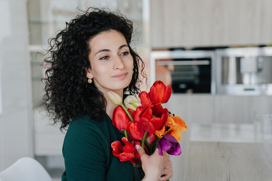 Pretty greatful curly italian young woman sits at table holds colourful tulips looks aside dreamily . Remote working hispanic businesswoman received flowers on women day, mother's day. Happy valentine
