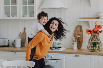 Cheerful son at kitchen riding  on moms back laughs happy spend time together. Curly Spanish woman...
