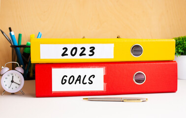 Red and yellow folders lie on the office table next to the clock and pen. Inscriptions on 2023 and GOALS folders.