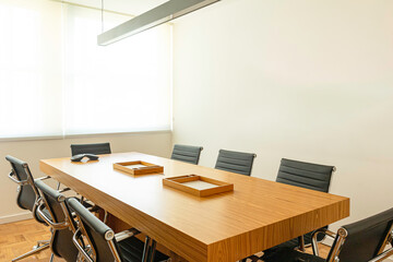 Close da mesa de reunião classica com papeleiras e aparelho de conferência, e ao redor cadeiras de couro preto ao fundo paredes e janelas com cortinas brancas. 
