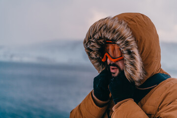 Headshot photo of a man in a cold snowy area wearing a thick brown winter jacket, snow goggles and...