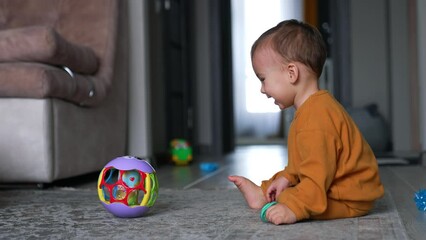 Happy laughing Caucasian baby boy playing jolly with a ball in the house. Smiling child kicks the ball ans watches it roll by the floor.