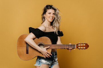 Pretty blonde with a cute smile in black top and jeans shorts holding acoustic guitar over yellow backdrop. Musicians practising at home. Hobbies for joy. Stay home during quarantine.