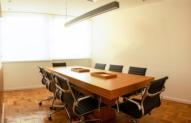 Sala de reunião e conferências clássica, com uma mesa de madeira com cadeiras de couro pretas ao seu redor, e ao fundo paredes brancas e uma janela. 