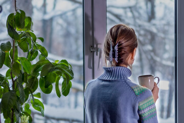 A woman in a blue sweater at the window with a snowy landscape is bored and drinking coffee....