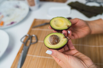 Hands of unrecognizable woman with an avocado cut in half ready to prepare homemade sushi in her kitchen.