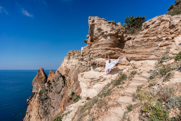 Fototapeta na wymiar A beautiful young woman in a white light dress with long legs stands on the edge of a cliff above the sea waving a white long dress, against the background of the blue sky and the sea.