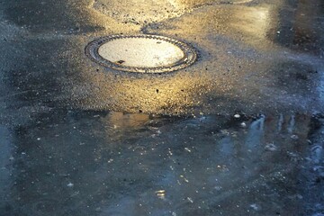 Nasse graue asphaltierte Straße mit Gullideckel und Regenpfütze mit Spiegelung bei Regen am Abend im Winter