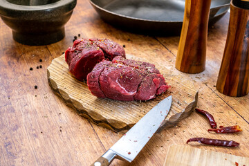 Preparation of raw beef fillet steak on wooden board with steel pan, mortar salt and pepper shakerand rosemary and chilly cutting board photographed from the side.