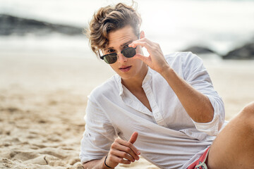 Portrait of handsome young italian man relaxing at the beach, wearing white shirt and fashionable sunglasses. - 555956005
