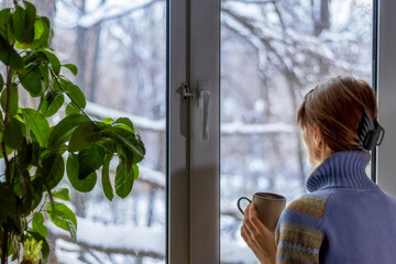 A woman in a blue sweater at the window with a snow-covered landscape drinks tea and looks...