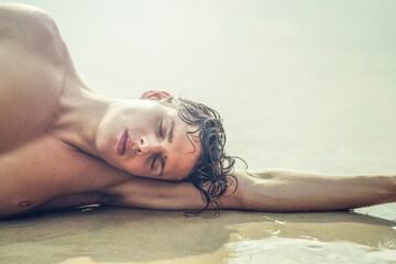 Beauty portrait of handsome young male model relaxing at the beach, lying on the sand. - 555956043