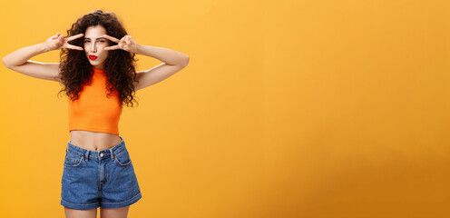 Portrait of good-looking daring and sexy stylish woman with curly hairstyle in red lipstick and cropped top showing victory or peace gestures overs eyes squinting folding lips flirty over orange wall